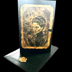 May include: A black greeting card featuring a sepia-toned portrait of a person with glowing white eyes and an elaborate updo hairstyle. The portrait has a distressed, torn-edge border. A black envelope with a gold wax seal sits in the foreground.