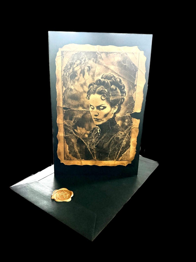 May include: A black greeting card featuring a sepia-toned portrait of a person with glowing white eyes and an elaborate updo hairstyle. The portrait has a distressed, torn-edge border. A black envelope with a gold wax seal sits in the foreground.