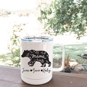 May include: White travel mug featuring a black bear design with floral patterns and the word "mama". The bear is walking, and below are the names Jenae, Javin, and Hailey with hearts. The mug has a white handle and a clear lid.