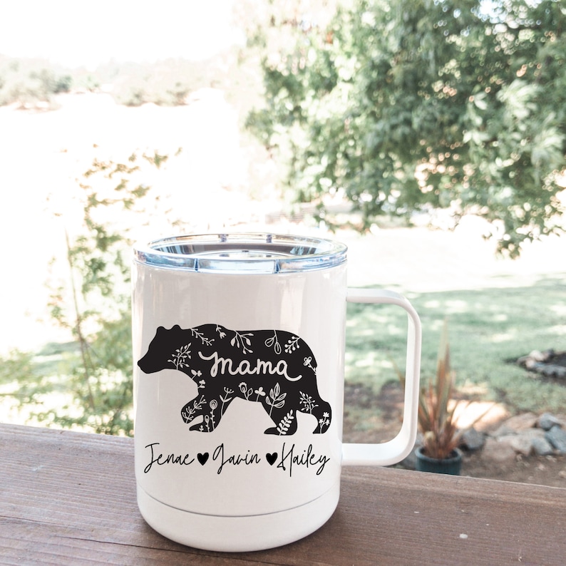 May include: White travel mug featuring a black bear design with floral patterns and the word "mama". The bear is walking, and below are the names Jenae, Javin, and Hailey with hearts. The mug has a white handle and a clear lid.