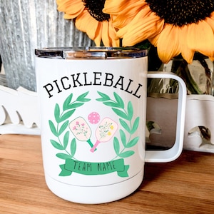May include: White stainless steel travel mug with a black and green design. The mug features a wreath with two pickleball paddles and a ball in the centre. The text "Pickleball" is at the top and "Team Name" is at the bottom.