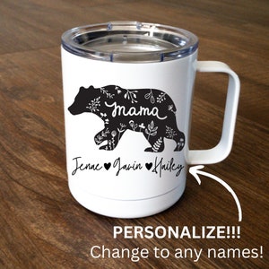 May include: White insulated travel mug featuring a black bear silhouette filled with floral designs. The word "mama" is inside the bear. Below the bear are the names "Jenae", "Javin", and "Hailey". The mug has a white handle and a clear lid.