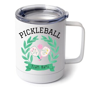 May include: White stainless steel travel mug with a black and green design. The design features a wreath of green leaves with two pickleball paddles and a ball inside. The text "Pickleball" is above the wreath and "Team Name" is below the wreath.