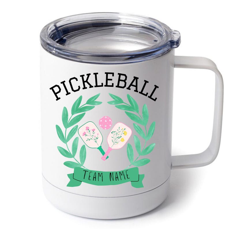 May include: White stainless steel travel mug with a black and green design. The design features a wreath of green leaves with two pickleball paddles and a ball inside. The text "Pickleball" is above the wreath and "Team Name" is below the wreath.