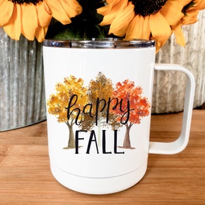 May include: White ceramic mug with a black handle. The mug has a design of three trees with fall foliage in shades of orange, yellow, and red. The text "happy fall" is printed in black below the trees.
