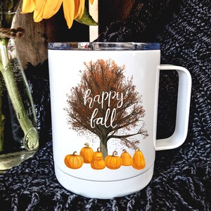 May include: White travel mug with a fall-themed design. The mug features a brown tree with orange leaves and the text "happy fall". There are six orange pumpkins at the base of the tree.