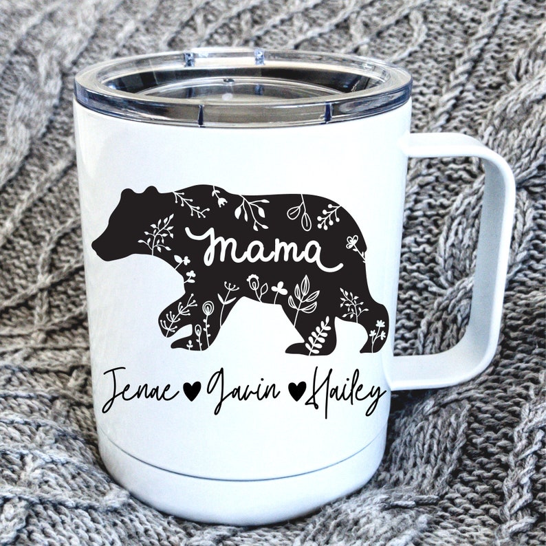 May include: White insulated coffee mug featuring a black bear silhouette filled with floral patterns. The word "mama" is in the center of the bear. Below the bear are the names "Jenae", "Javin", and "Hailey" with a heart symbol between each name.