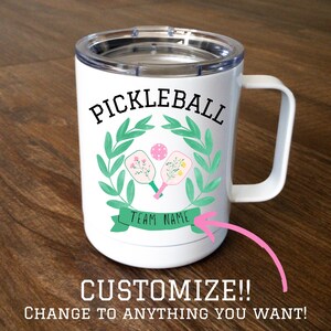 May include: White stainless steel travel mug with a lid. The mug has a green wreath with two pink and yellow pickleball paddles and a pink ball inside. The text "Pickleball" is above the wreath and "Team Name" is below the wreath.