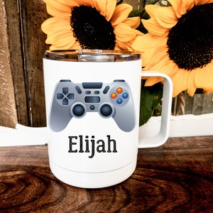May include: White travel mug with a gray video game controller graphic and the name Elijah printed in black.