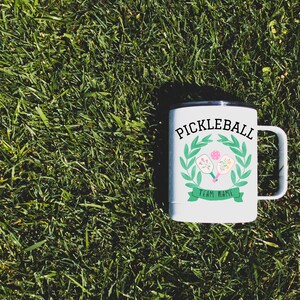 May include: White travel mug with a black lid and handle. The mug features a green wreath with two pink and white pickleball paddles decorated with flowers. The text "Pickleball" is in black letters above the wreath and "Team Name" is in black letters below the wreath.