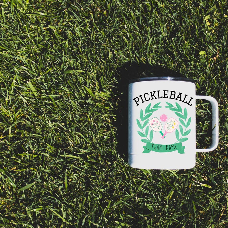 May include: White travel mug with a black lid and handle. The mug features a green wreath with two pink and white pickleball paddles decorated with flowers. The text "Pickleball" is in black letters above the wreath and "Team Name" is in black letters below the wreath.