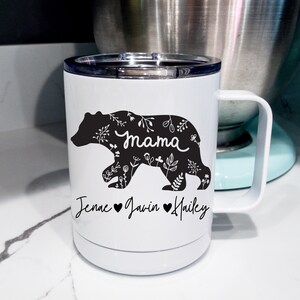 May include: White insulated travel mug featuring a black bear silhouette filled with floral patterns. The word "Mama" is written in cursive within the bear. Below the bear are the names "Jenae", "Javin", and "Hailey" with hearts.