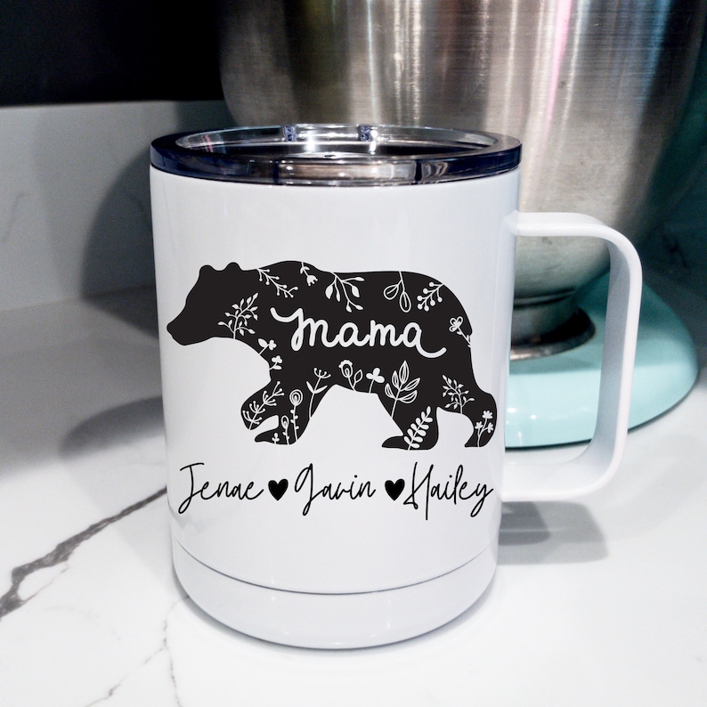 May include: White insulated travel mug featuring a black bear silhouette filled with floral patterns. The word "Mama" is written in cursive within the bear. Below the bear are the names "Jenae", "Javin", and "Hailey" with hearts.