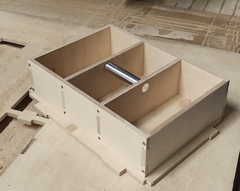Op de afbeelding: Een close-up van een houten doos met compartimenten die worden gesneden door een CNC-machine. De machine heeft een blauw en zwart lichaam met de tekst "S H300" aan de voorkant. De doos heeft een metalen staaf in een van de compartimenten.