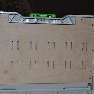 May include: A light-colored wooden panel with evenly spaced rectangular cutouts, set within a gray plastic frame. The panel is part of a larger storage or organizational system, with a green handle visible at the top.