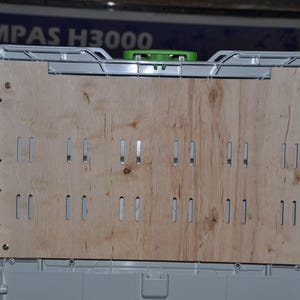 May include: A light gray storage container with a light brown wooden panel. The panel has multiple rectangular cutouts. The container has a metal frame and is secured with screws. The text "MPAS H3000" is visible in the background.