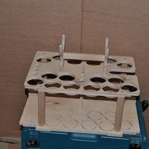 Puede incluir: Un organizador de botellas de pintura de madera con múltiples agujeros circulares para sujetar botellas. El organizador está hecho de madera de color claro y tiene un asa y soportes verticales. Está diseñado para colocarse encima de un contenedor de almacenamiento turquesa.