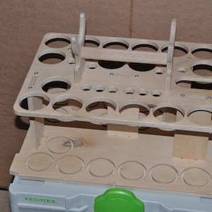 Puede incluir: Un organizador de herramientas de madera de dos niveles con numerosos recortes circulares de diferentes tamaños. El organizador de madera de color claro está diseñado para guardar herramientas. Se encuentra sobre un contenedor gris y verde.