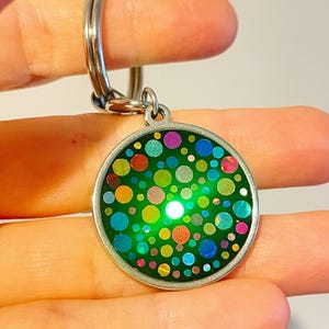 May include: A circular keyring pendant featuring a green base adorned with multi-coloured polka dots. The pendant is framed in silver-coloured metal and has a silver key ring.