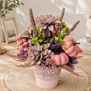 Könnte beinhalten: Ein Blumenarrangement in einem hellrosa Metalltopf mit dekorativem Ausschnittmuster. Der Strauß besteht aus staubig malvenfarbenen, dahlienartigen Blüten, zwei kleinen hellrosa Kürbissen, rosa Beerenclustern, violetten Zweigen und bronzefarbenen Farnblättern.