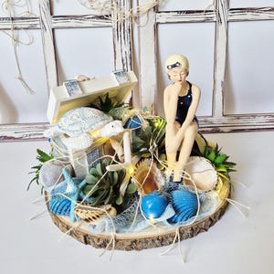 Könnte beinhalten: Dekorative Tischdekoration zum Thema Strand auf Holzbasis. Sie zeigt eine Figur einer Frau im Badeanzug, Muscheln, eine Schatzkiste, eine Schildkröte und blaue Seesterne. Die Anordnung umfasst Grün und Lichterketten, wodurch eine Küstenästhetik entsteht.