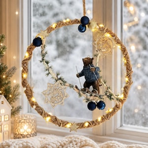 Könnte beinhalten: Ein kreisförmiger Hängekranz aus gedrehtem Naturseil, beleuchtet durch kleine warme LED-Lichter. Der Kranz ist mit dunkelblauen kugelförmigen Ornamenten, goldenen Glitzersternen und einer zentralen Figur eines braunen Bären in blauem Pullover verziert.