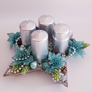 Könnte beinhalten: Ein silberner Adventskranz mit vier silbernen Kerzen. Die sternförmige Basis ist mit türkisfarbenen Blumen, silbernen Perlen und grünen Akzenten verziert. Ein festlicher Mittelpunkt für die Weihnachtszeit.