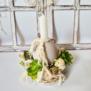 Könnte beinhalten: Ein dekorativer Kerzenhalter mit einer beigefarbenen Keramikvase, die eine weiße Kerze hält. Die Vase ist mit Seilen verziert und von künstlichen Sukkulenten, Muscheln und einer kleinen Schildkrötenfigur umgeben. Der Sockel hat ein dekoratives Muster.