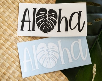 May include: Two vinyl stickers with the word "Aloha" in a cursive font. The word is centered on each sticker with a black monstera leaf in the center of the word on the white sticker and a white monstera leaf in the center of the word on the blue sticker.