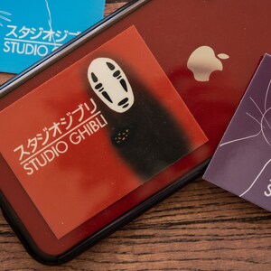 Op de afbeelding: Een rode telefoonhoes met een wit No Face-personage van Studio Ghibli. De hoes heeft een zwarte rand en een wit Apple-logo. Er liggen drie Studio Ghibli-stickers op de tafel.