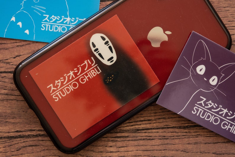 Op de afbeelding: Een rode telefoonhoes met een wit No Face-personage van Studio Ghibli. De hoes heeft een zwarte rand en een wit Apple-logo. Er liggen drie Studio Ghibli-stickers op de tafel.