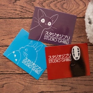 Op de afbeelding: Drie Studio Ghibli stickers op een houten oppervlak. De stickers tonen een kat, een personage zonder gezicht en Totoro. De stickers zijn wit op een paarse, rode en blauwe achtergrond.