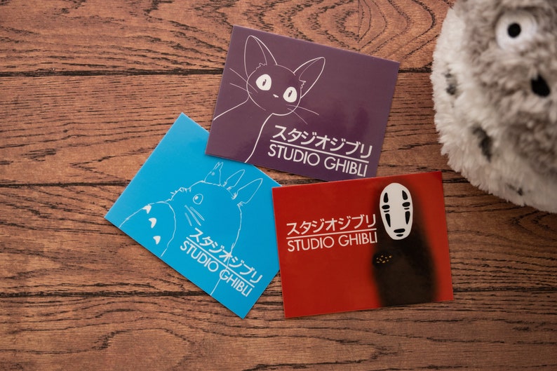 Op de afbeelding: Drie Studio Ghibli stickers op een houten oppervlak. De stickers tonen een kat, een personage zonder gezicht en Totoro. De stickers zijn wit op een paarse, rode en blauwe achtergrond.
