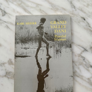 May include: A book cover with a black and white photo of a person walking through water. The book title is "Grand Valley Dani: Peaceful Warriors" and the author is Karl Heider.