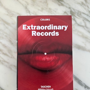 Puede incluir: Un libro rojo titulado "Extraordinary Records" con la palabra "COLORS" en la parte superior. La portada presenta un primer plano de un diseño de disco de vinilo rojo. El nombre del editor, TASCHEN, está en la parte inferior.