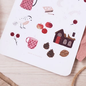 Puede incluir: Una hoja de pegatinas con ilustraciones de tem&aacute;tica invernal, que incluyen un p&aacute;jaro rojo, una taza roja, una casa roja con nieve en el techo y manzanas rojas. Las pegatinas est&aacute;n sobre un fondo blanco.