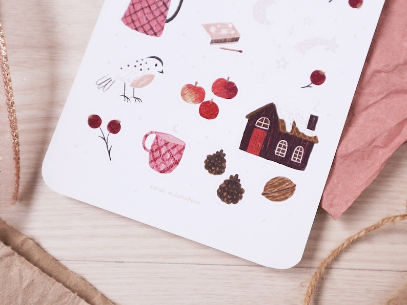 Puede incluir: Una hoja de pegatinas con ilustraciones de tem&aacute;tica invernal, que incluyen un p&aacute;jaro rojo, una taza roja, una casa roja con nieve en el techo y manzanas rojas. Las pegatinas est&aacute;n sobre un fondo blanco.