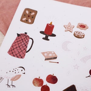 Puede incluir: Una hoja de pegatinas con ilustraciones de art&iacute;culos con tem&aacute;tica navide&ntilde;a de invierno, que incluyen una vela roja, una tetera, galletas de jengibre, un p&aacute;jaro, manzanas y una estrella. Las pegatinas est&aacute;n sobre un fondo blanco con un sutil patr&oacute;n rosa.
