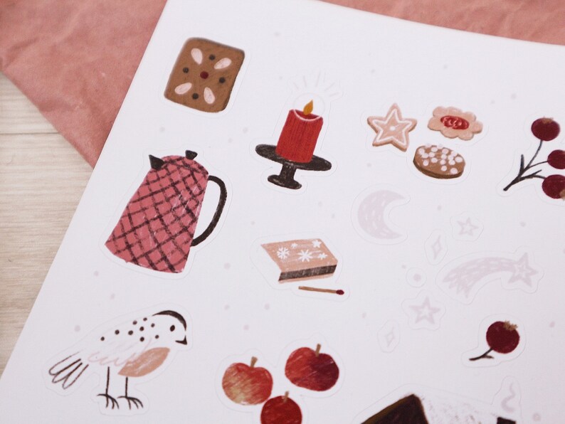 Puede incluir: Una hoja de pegatinas con ilustraciones de art&iacute;culos con tem&aacute;tica navide&ntilde;a de invierno, que incluyen una vela roja, una tetera, galletas de jengibre, un p&aacute;jaro, manzanas y una estrella. Las pegatinas est&aacute;n sobre un fondo blanco con un sutil patr&oacute;n rosa.