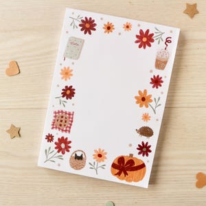 Könnte beinhalten: Ein weißer Notizblock mit einem floralen Rand mit roten und orangefarbenen Blumen, einem Kürbis und einer Kaffeegetränke-Illustration. Dekorative Holzherzen und -sterne sind um den Notizblock auf einer hellen Holzoberfläche verstreut.