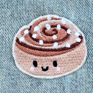 Könnte beinhalten: Gestickter Aufnäher mit einem lächelnden Zimtschnecken-Design. Der Aufnäher hat eine hellbraune Basis mit dunkelbraunen Wirbeln und weißen Streuseln. Die Zimtschnecke hat ein süßes Gesicht mit schwarzen Augen und einem lächelnden Mund. Der Aufnäher befindet sich auf einem blauen Jeans-Hintergrund.