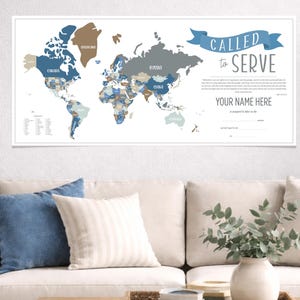 May include: A framed world map with countries in shades of blue, brown, and gray. The text "CALLED to SERVE" is above a space for a name. The map is displayed above a beige sofa with blue and white pillows.
