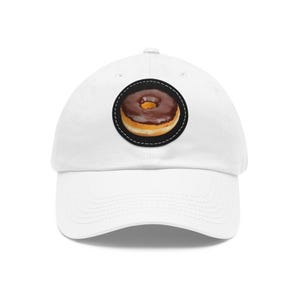 Puede incluir: Una gorra de béisbol blanca con un parche circular en el panel frontal. El parche muestra una imagen realista de una dona redonda con glaseado de chocolate marrón oscuro y una base de masa clara, rodeada por un borde negro con costuras blancas.
