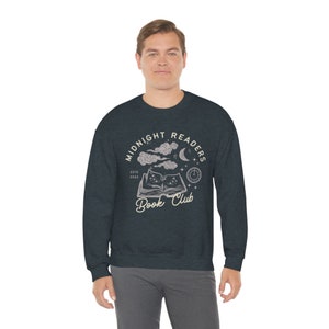 May include: A dark blue sweatshirt with a white graphic design of a book with stars, clouds, a crescent moon, and a clock. The text on the graphic reads "Midnight Readers Book Club Est. 2023".
