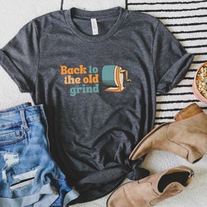 以下が含まれることがあります： オレンジとティール色の「Back to the old grind」の文字が入った濃いグレーのTシャツ。ヴィンテージの鉛筆削りのグラフィック。デニムショーツとタンカラーのアンクルブーツが組み合わされています。