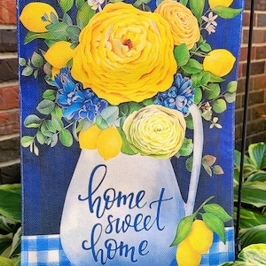 May include: A decorative garden flag featuring a blue background with a white pitcher filled with yellow roses, lemons, and blue flowers. The words "home sweet home" are written in cursive. A blue and white checkered pattern is at the bottom.
