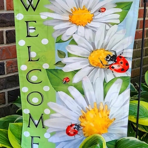 May include: A rectangular garden flag featuring the word WELCOME in black capital letters on a vertical lime green stripe with white polka dots. The main design displays three large white daisies with yellow centers and several red ladybugs.
