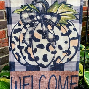 May include: A rectangular garden flag featuring a central pumpkin with a leopard print pattern, accented by a black and white buffalo check bow and green leaves. The background has a blue and white plaid pattern, with the word WELCOME in blue.