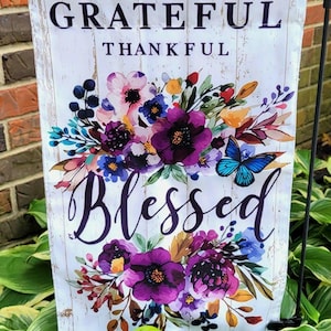 May include: A white garden flag with the words "Grateful, Thankful, Blessed" in black script. The flag features a floral design with purple, pink, blue, and yellow flowers, and a blue butterfly. The background has a wood-like texture.