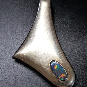 Könnte beinhalten: Silberfarbener Anhänger in Tropfenform mit einem ovalen Opalstein. Der Opal zeigt ein Farbspektrum, darunter Grün, Blau und Orange. Der Anhänger hat eine glatte, polierte Oberfläche.